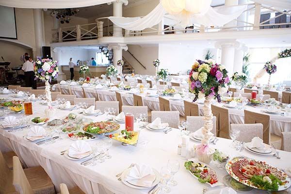A bright, elegant banquet hall set for a wedding with long white-clothed tables, floral centerpieces, and place settings.
