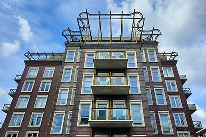 A low-angle view of a brick apartment building with white-framed windows, central balconies, and a metal roof structure.