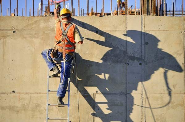 A construction worker in a high-visibility vest and safety harness climbs a metal ladder against a concrete wall.
