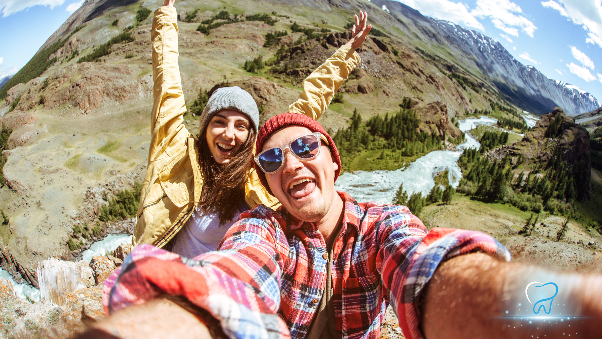 Two people smiling, taking a selfie on a mountain; green and grey mountains, river, sunny.