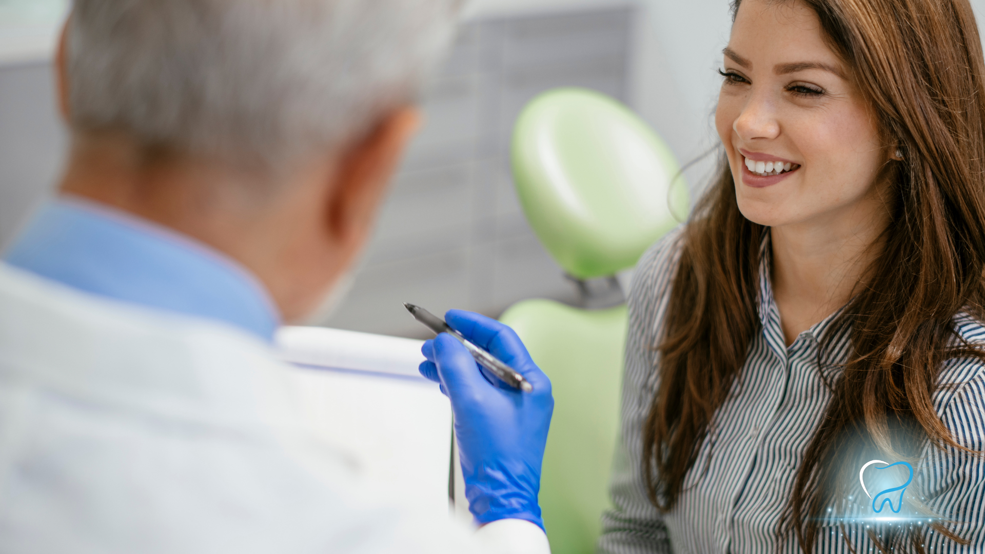 Dentist in gloves pointing pen, talking to smiling patient in office with green chair.