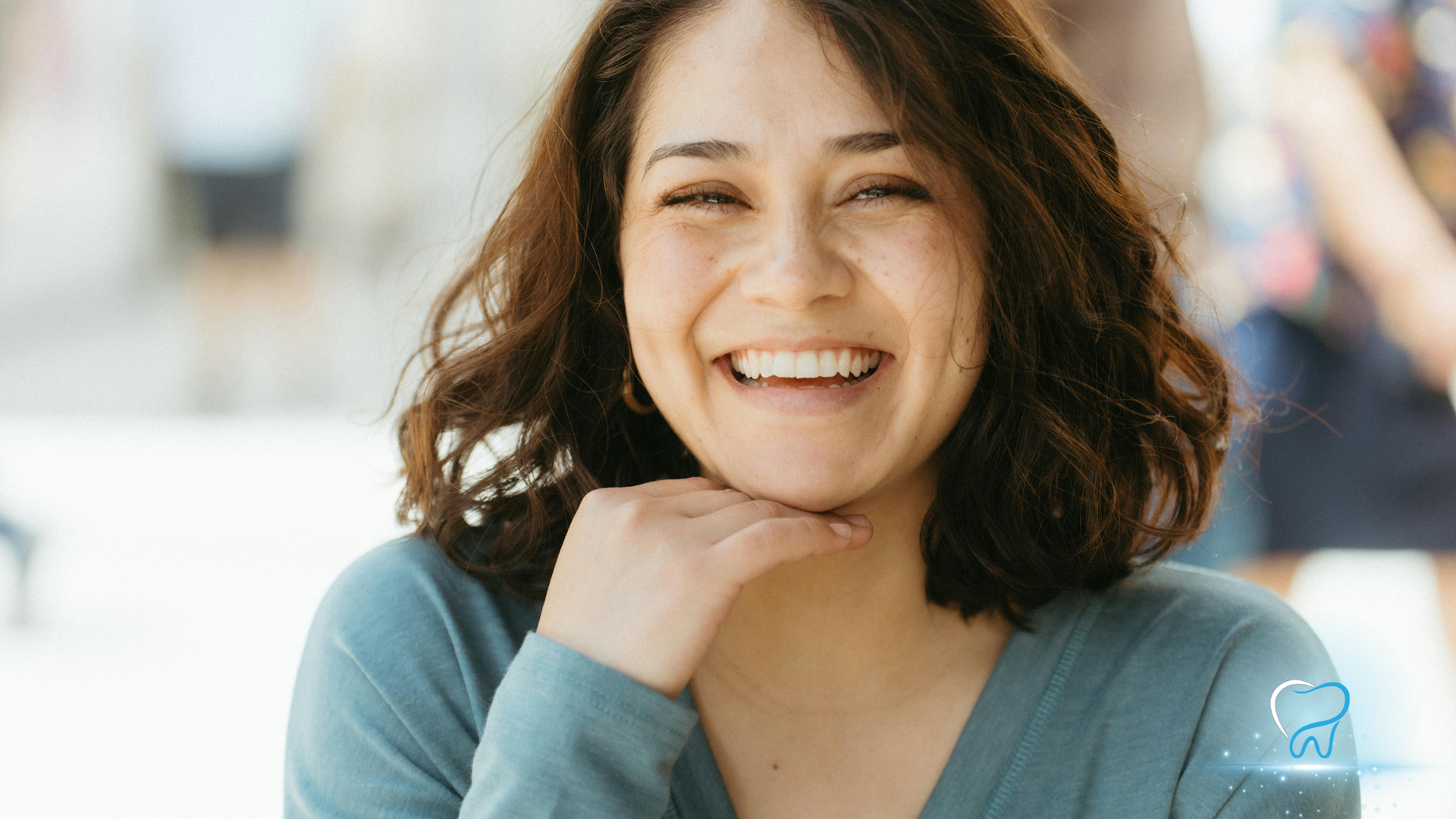 Woman smiles broadly, hand under chin. She wears a blue shirt outdoors in a bright setting.