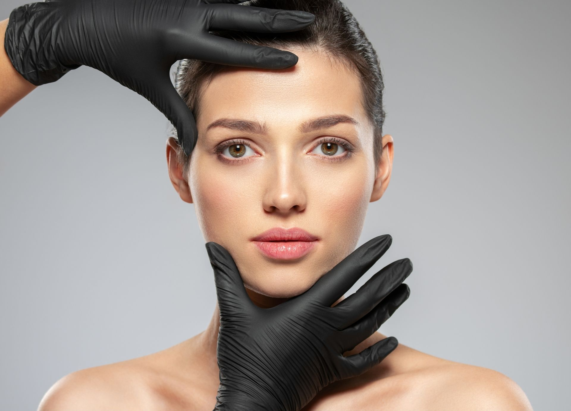 Close-up of a woman’s face being framed by black-gloved hands against a gray background