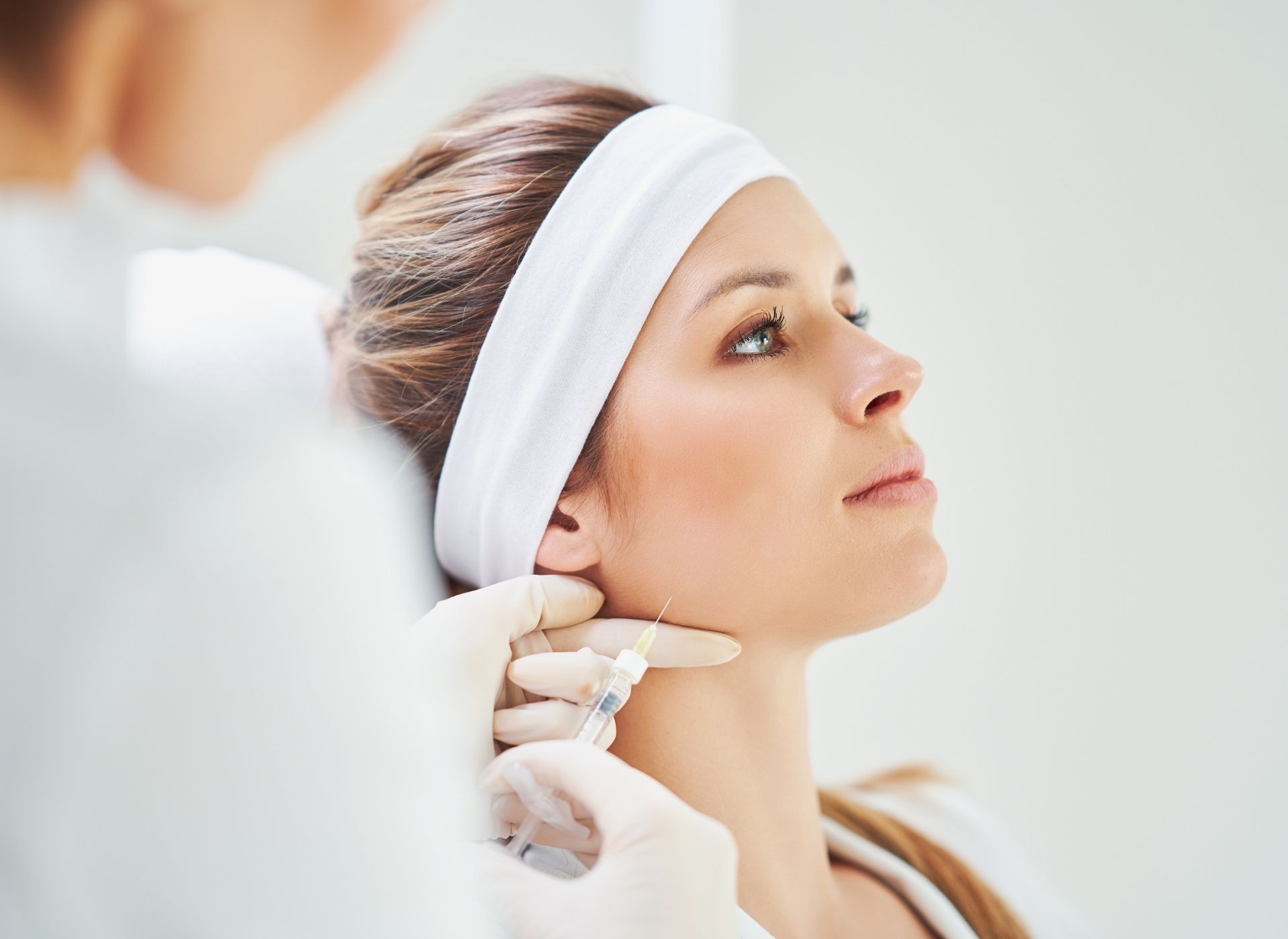 A cosmetic procedure on a woman’s jawline as a clinician wearing gloves works in a bright clinic