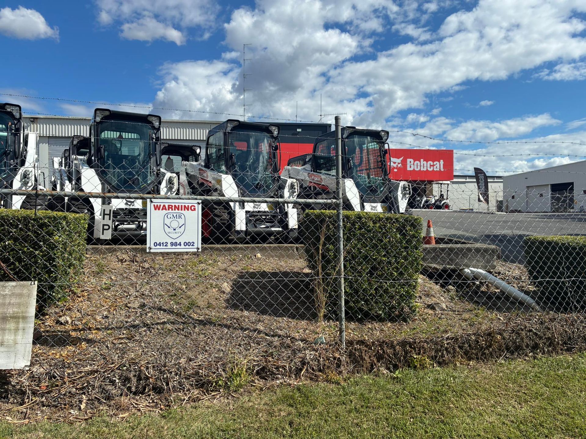Bobcat construction vehicles parked behind a chain-link fence in front of a building on a sunny day.