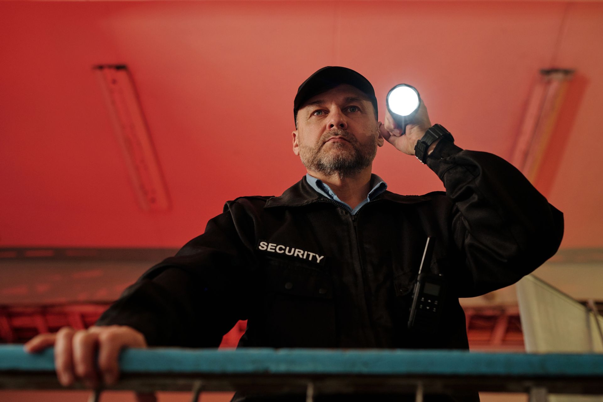 Security guard shining a flashlight. Wearing a cap, black jacket, looking vigilantly. Red background.