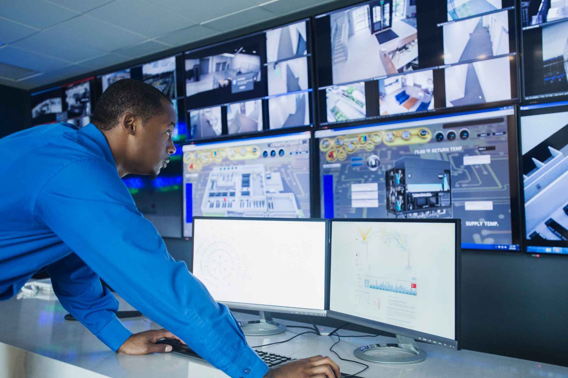 Man in blue shirt monitors security cameras in control room.