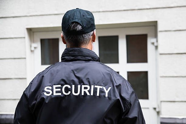 Security guard in black jacket and cap, facing a windowed door, with 