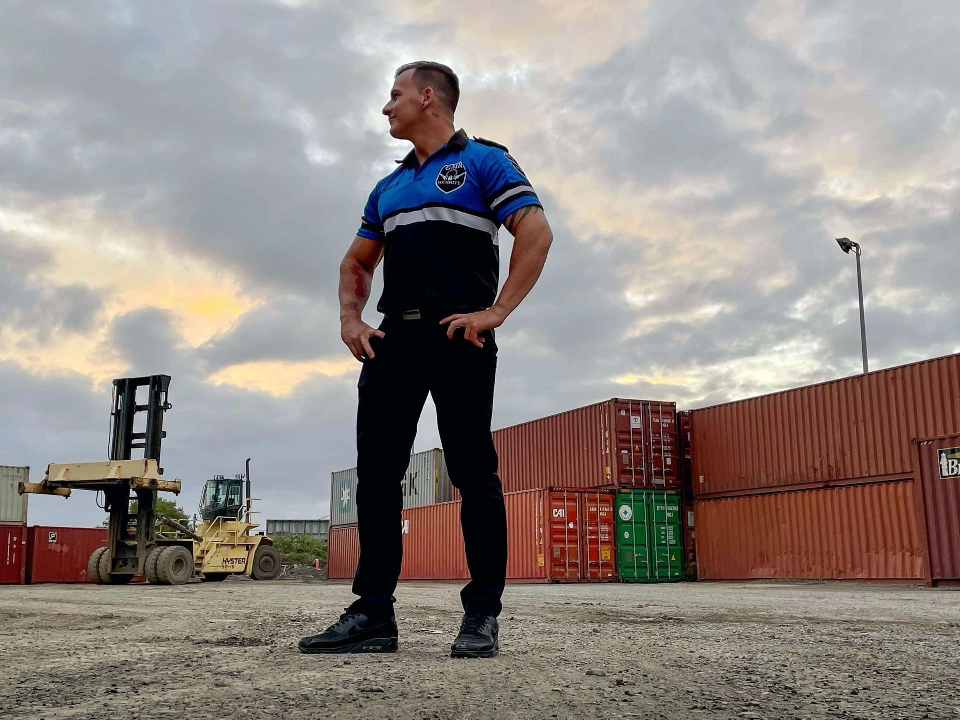 Man in blue and black shirt stands at shipping containers; looking off-camera. Cloudy sky.
