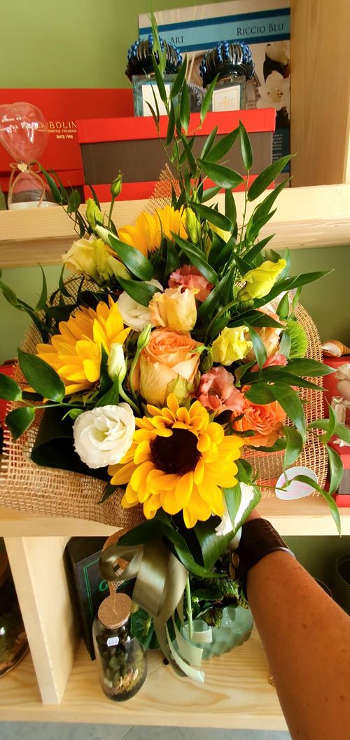 Una composizione floreale colorata con girasoli, rose e verde, in un cestino di vimini su uno scaffale.