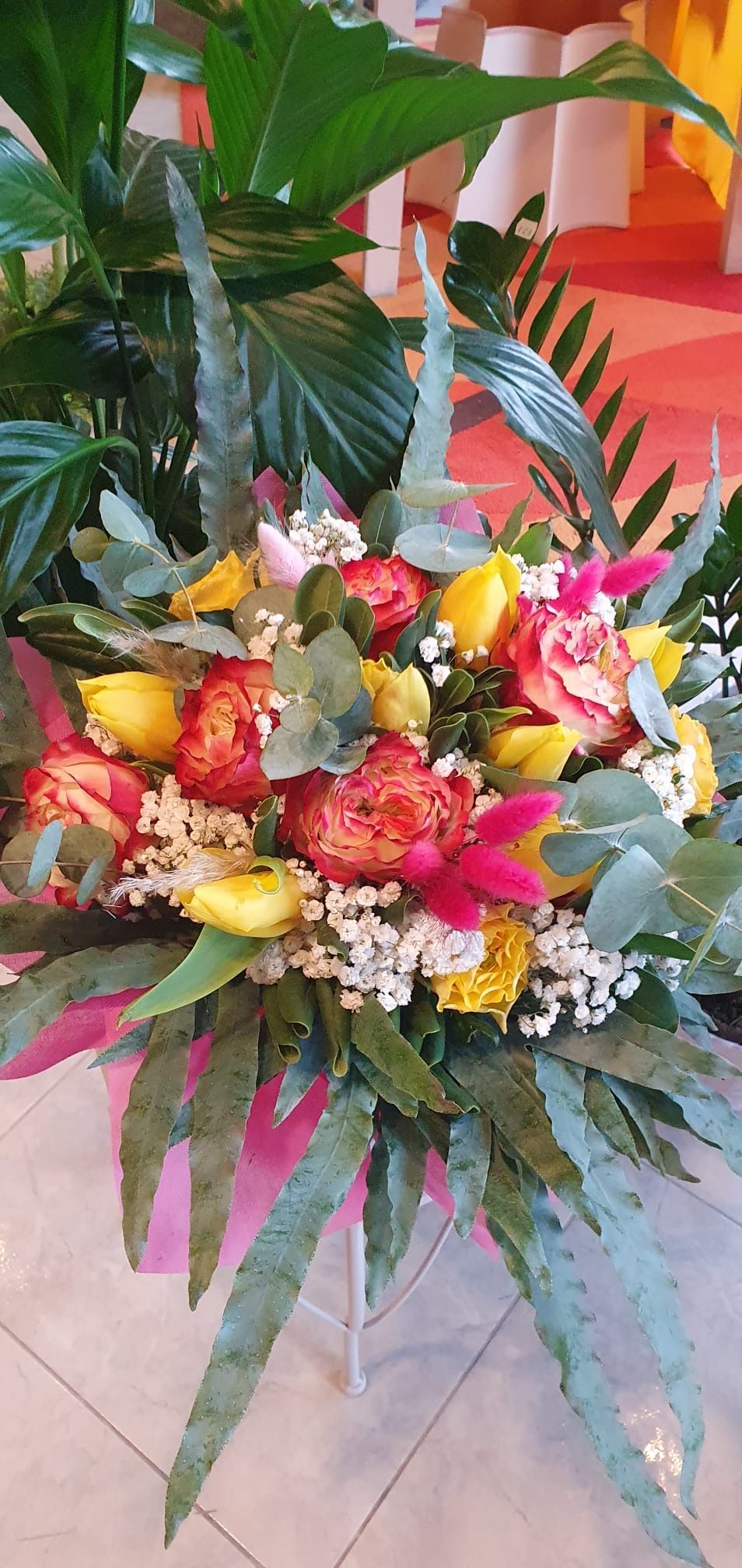 Mazzo di fiori gialli, rosa e rossi con fogliame verde, in un contenitore rosa.