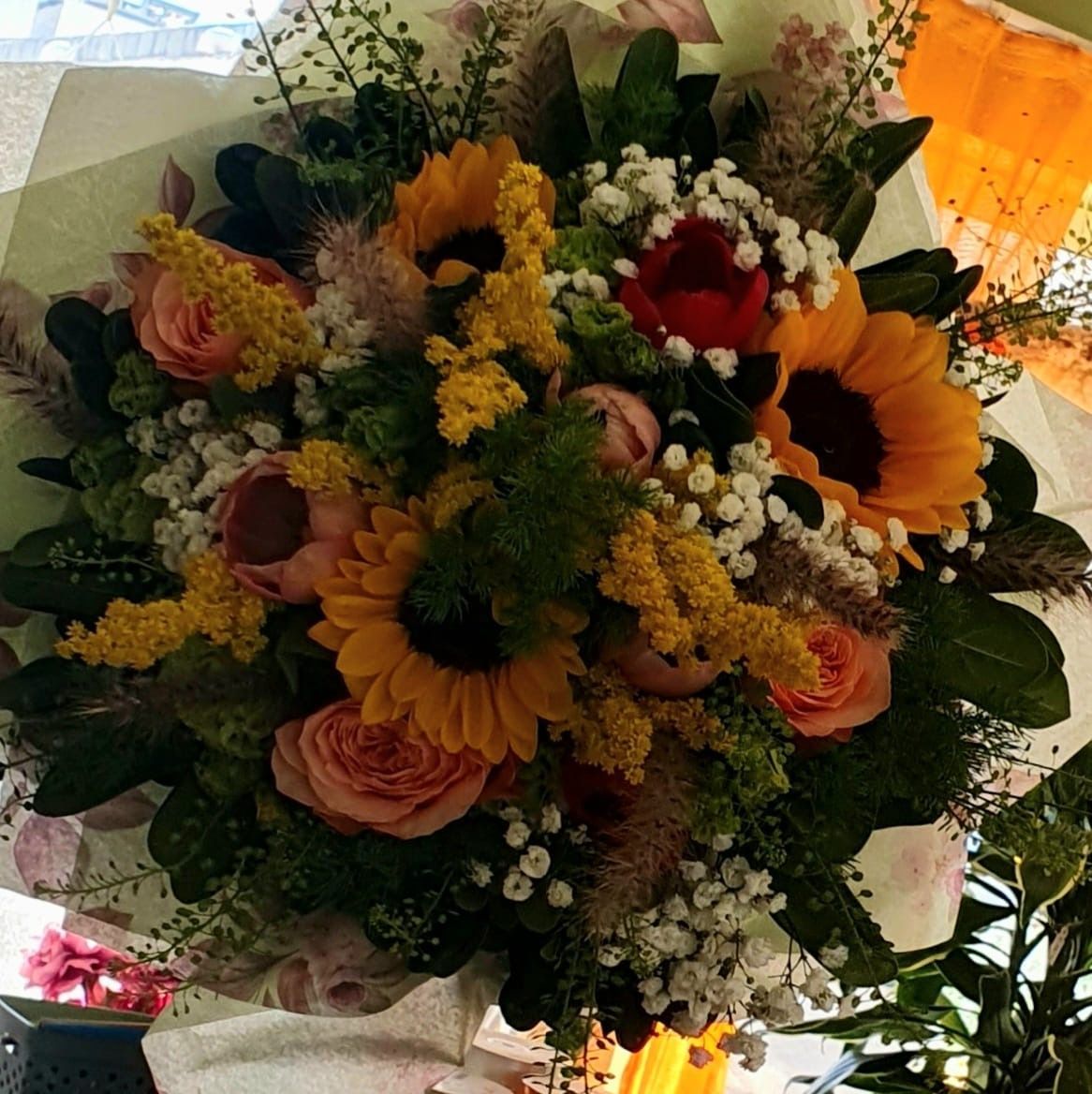 Bouquet di girasoli, rose e verde, avvolto in carta leggera, dall'aspetto luminoso e colorato.