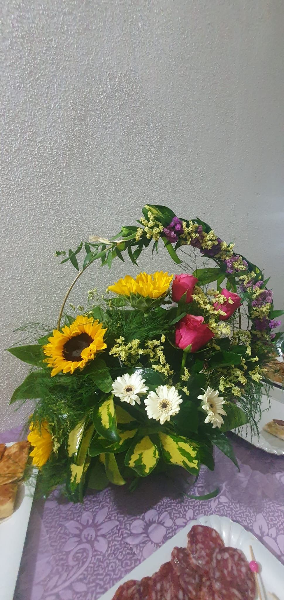 Composizione floreale con girasoli, rose e margherite su un tavolo con del cibo.