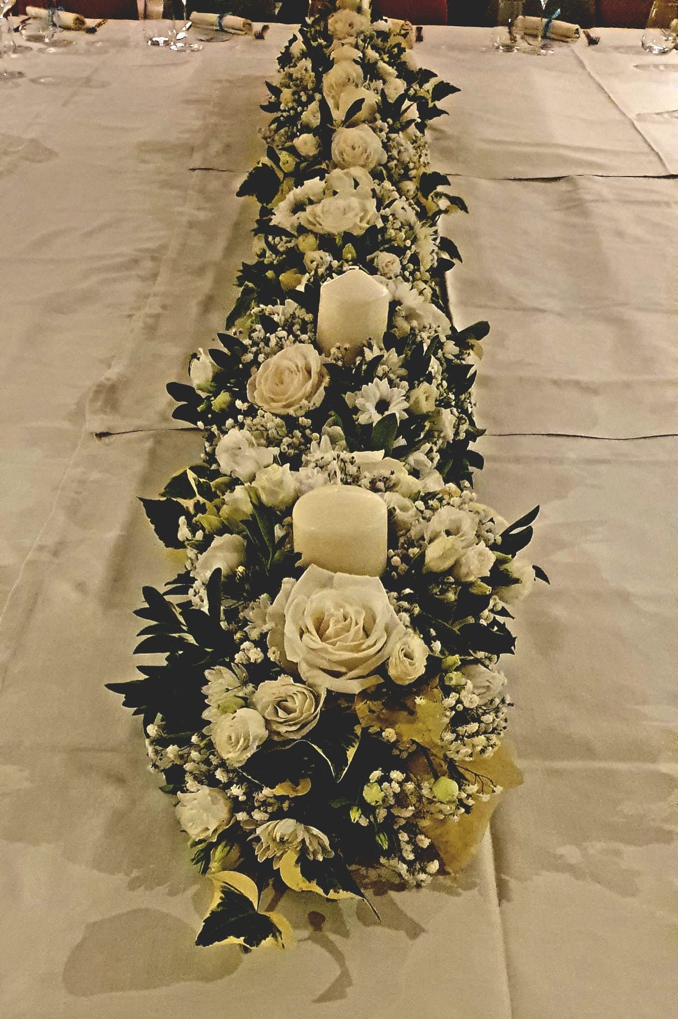 Runner da tavolo floreale con rose bianche, candele e verde su una tovaglia bianca.