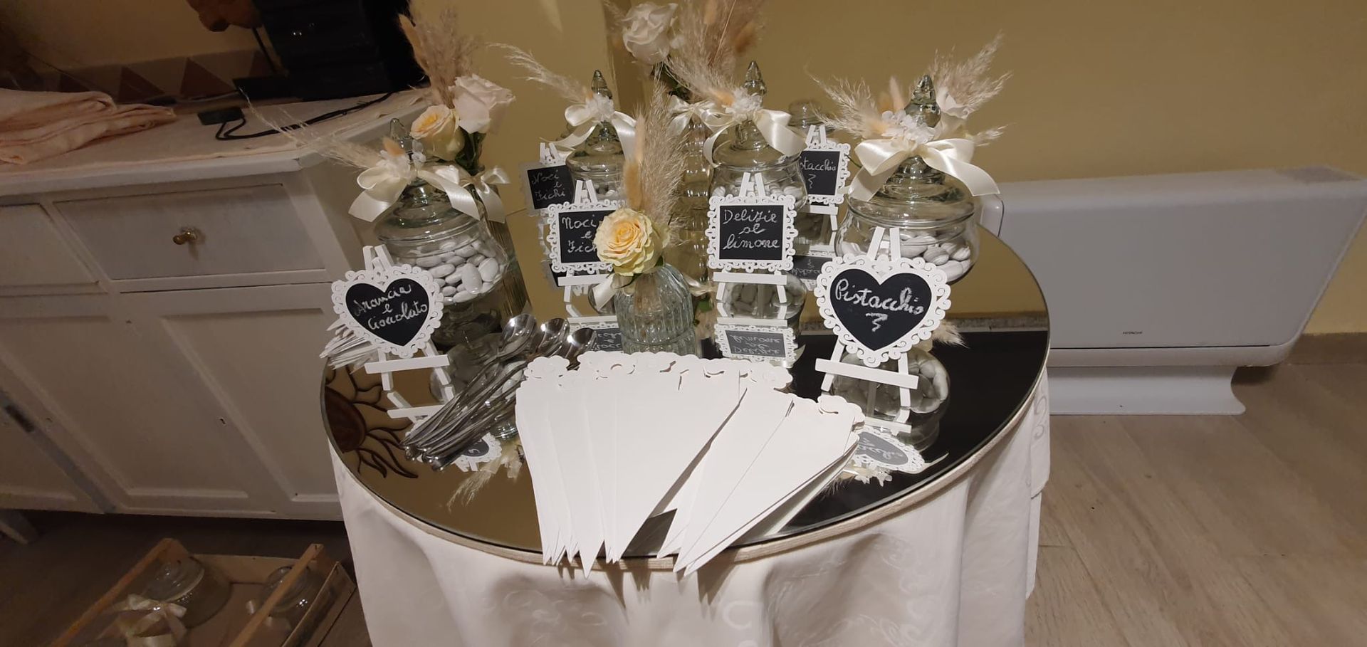 Un'esposizione di bomboniere su un tavolo rotondo a specchio, decorato con fiori e piccole lavagne.
