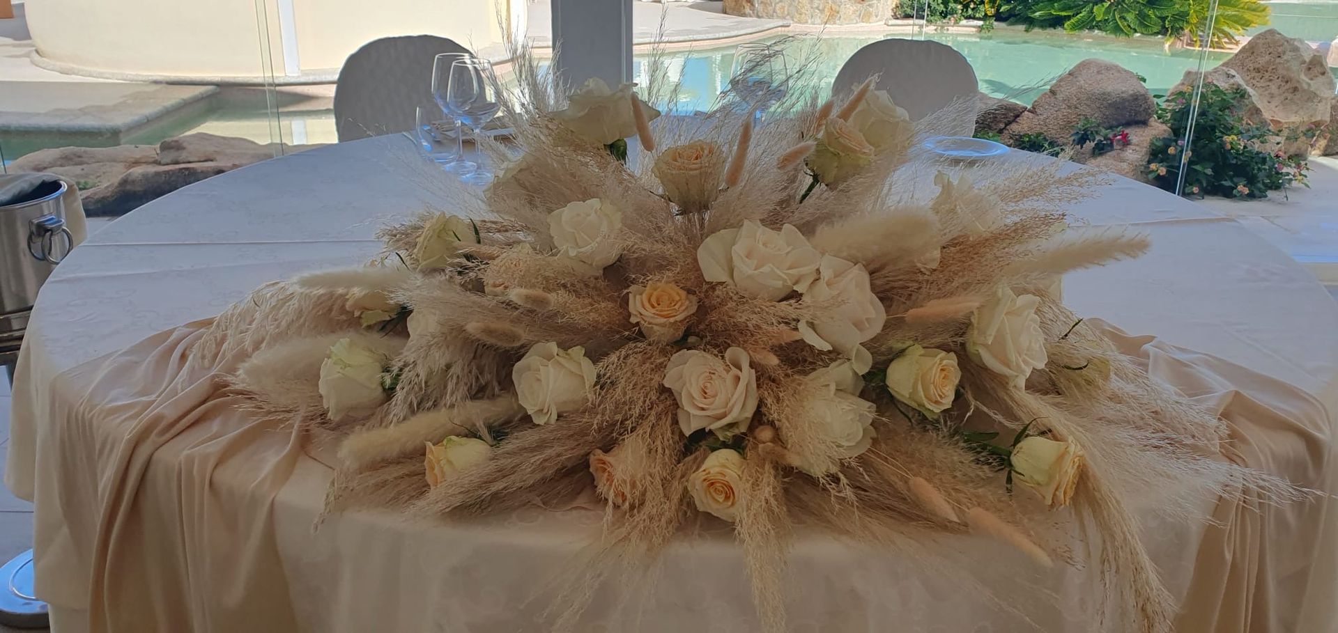 Un tavolo decorato con fiori bianchi ed erba della pampa, apparecchiato all'aperto vicino a una piscina.