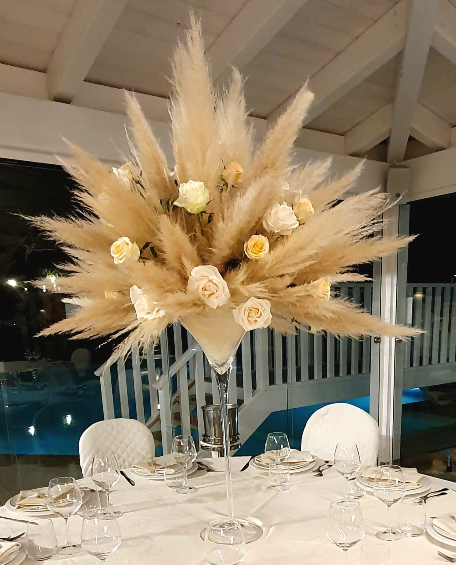 Centrotavola floreale con erba della pampa color crema e rose su un tavolo apparecchiato. Colori bianco e beige.