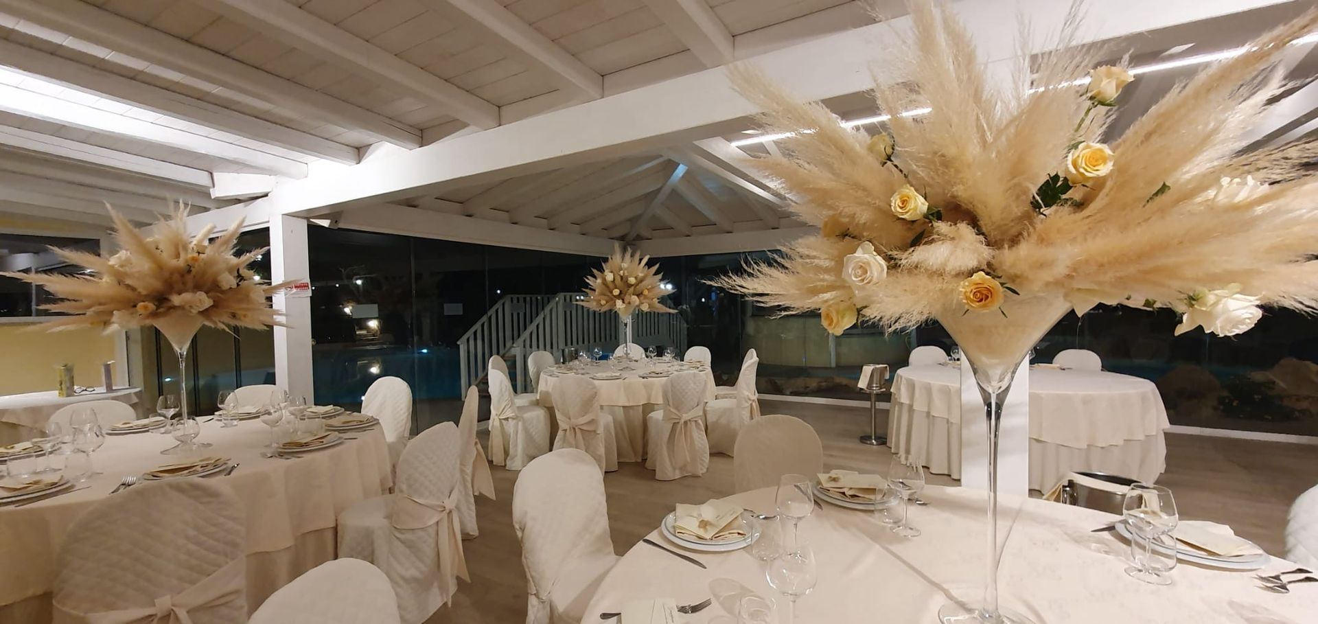 Una sala ricevimenti allestita per una festa. Tavoli drappeggiati di bianco, composizioni floreali.