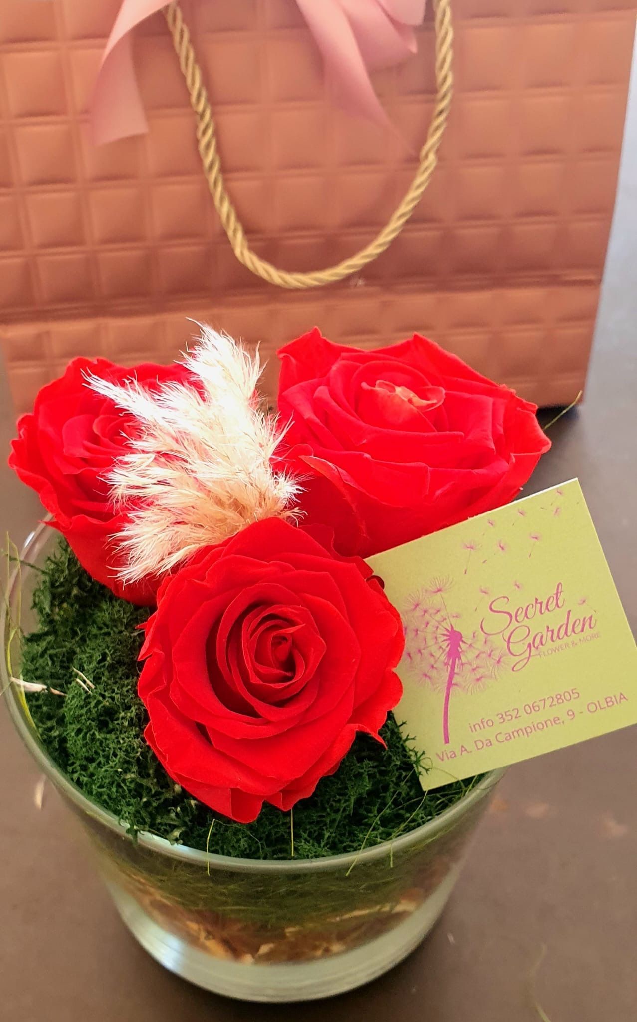 Tre rose rosse e una soffice pianta bianca in un bicchiere, con una borsa regalo dietro e un piccolo biglietto.