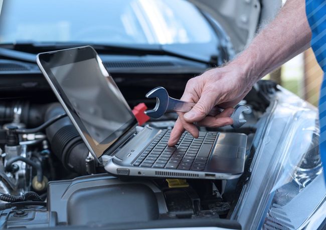 A man is working on a laptop computer under the hood of a car.