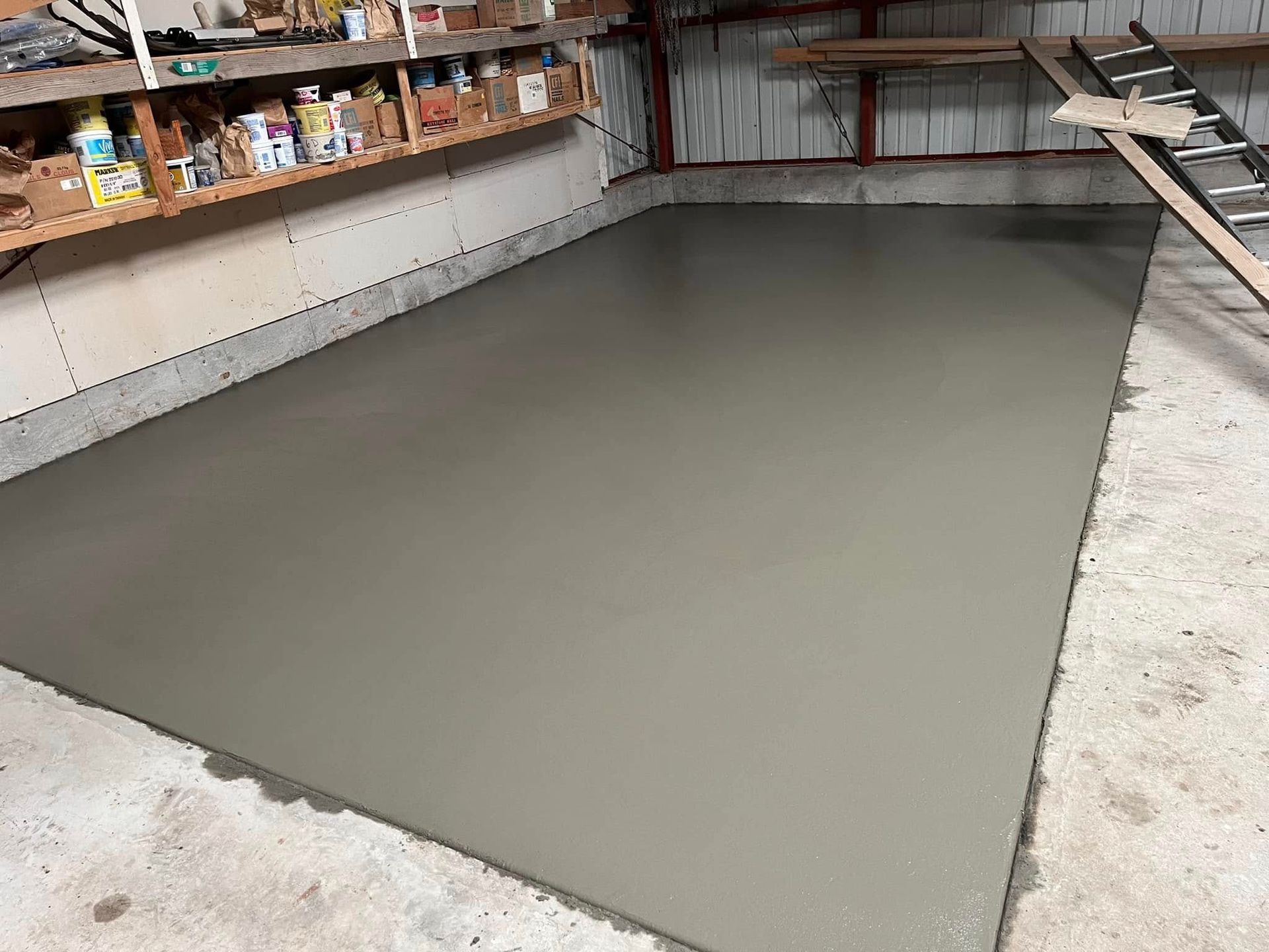 A smooth, freshly poured concrete floor section inside a garage with shelving units and a ladder in the background.