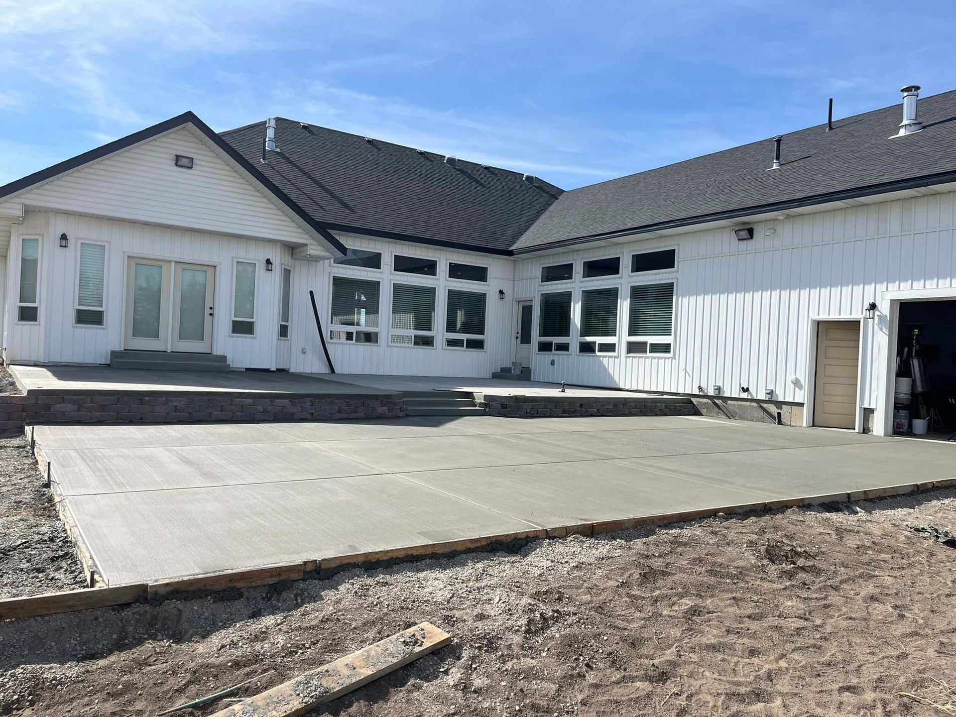 A wide view of a modern white house with a newly poured concrete patio and dark shingled roof under a clear blue sky.