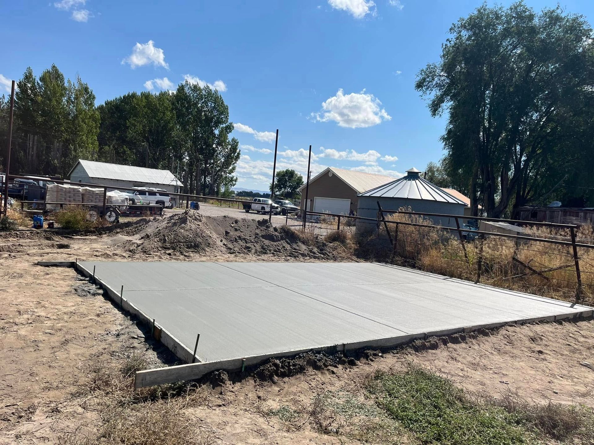 A freshly poured concrete slab sits in an outdoor, rural area, with buildings and trees visible in the background.