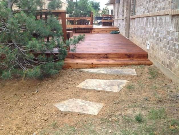 A wooden deck attached to a brick house, featuring three stone pavers leading across a dirt yard to the deck steps.
