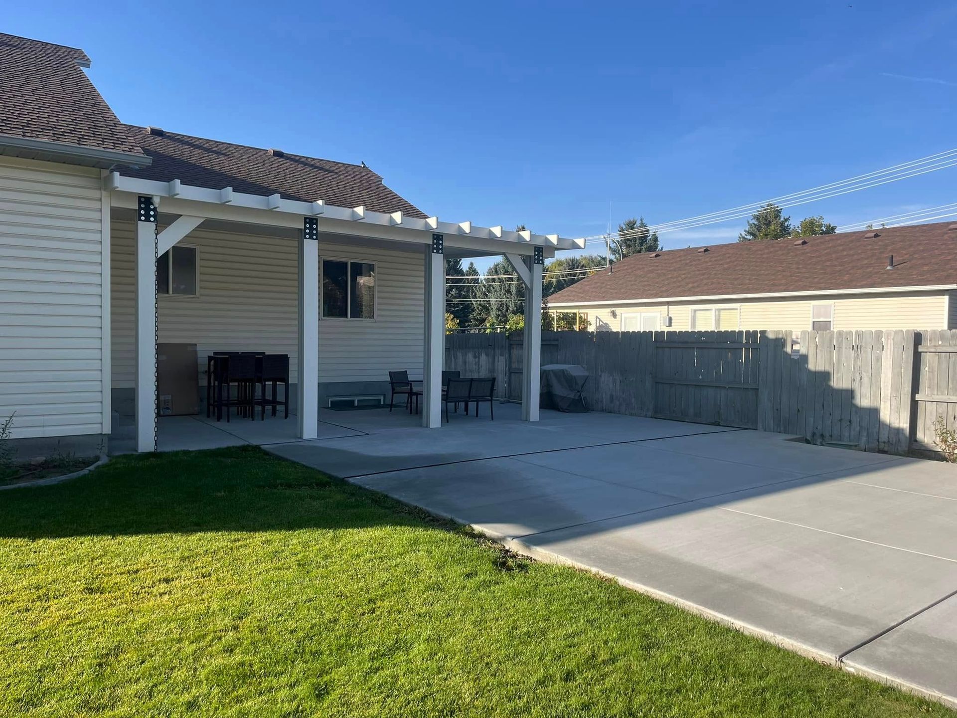 A beige house with a covered patio, a concrete slab, and a green lawn under a clear blue sky.