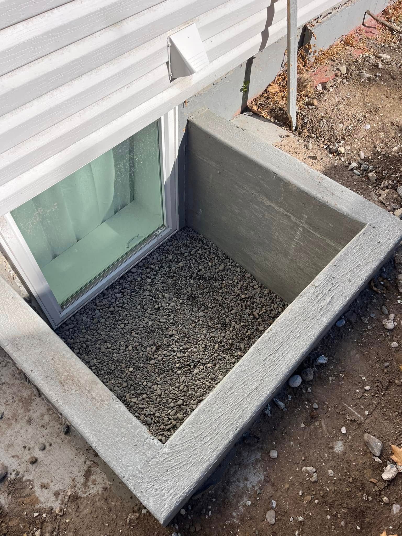 A rectangular window well with gray concrete walls, filled with gravel, installed against a white-sided house foundation.
