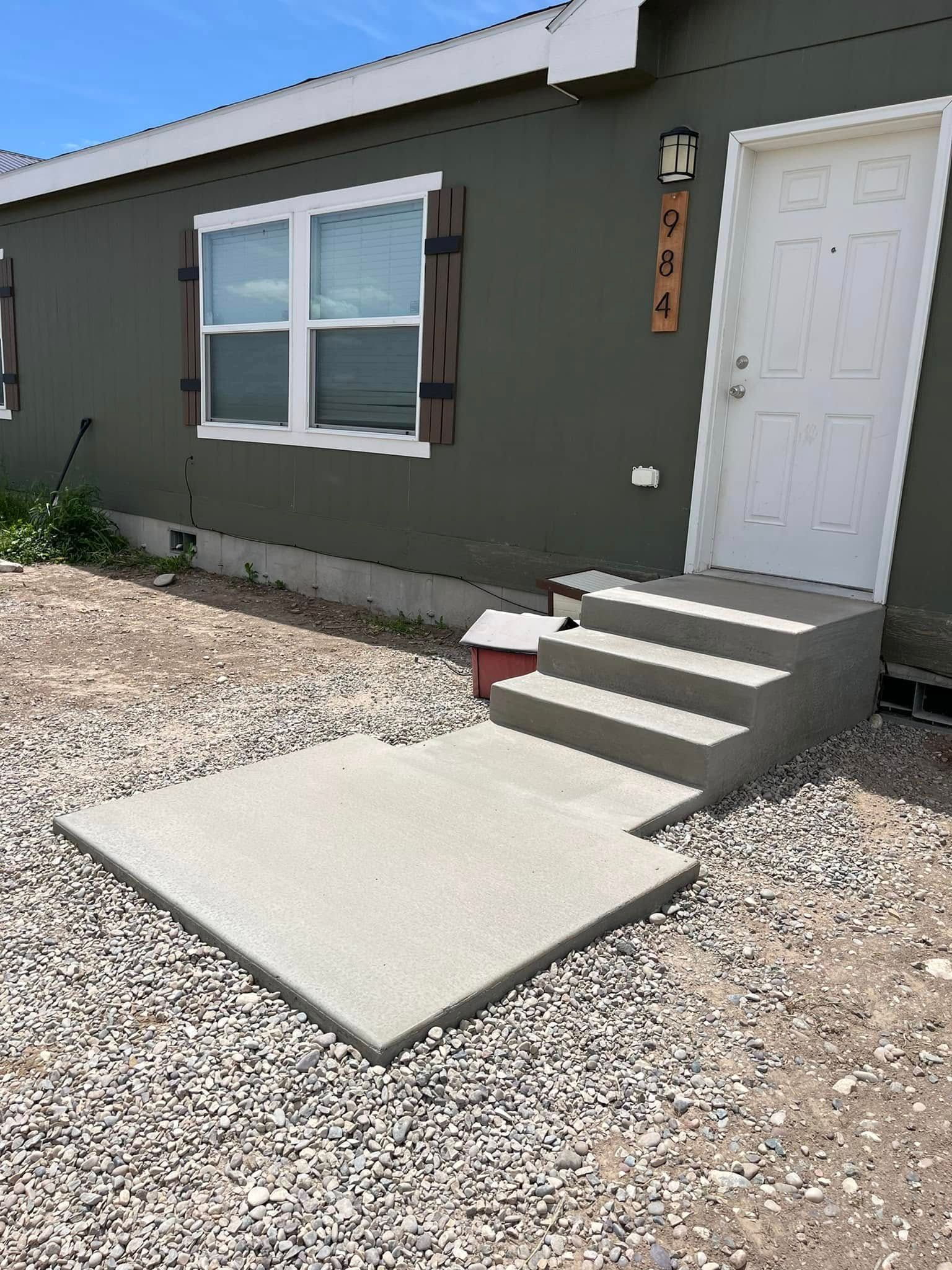 A green house with a white door and a new concrete landing with four steps leading to the entrance over gravel ground.