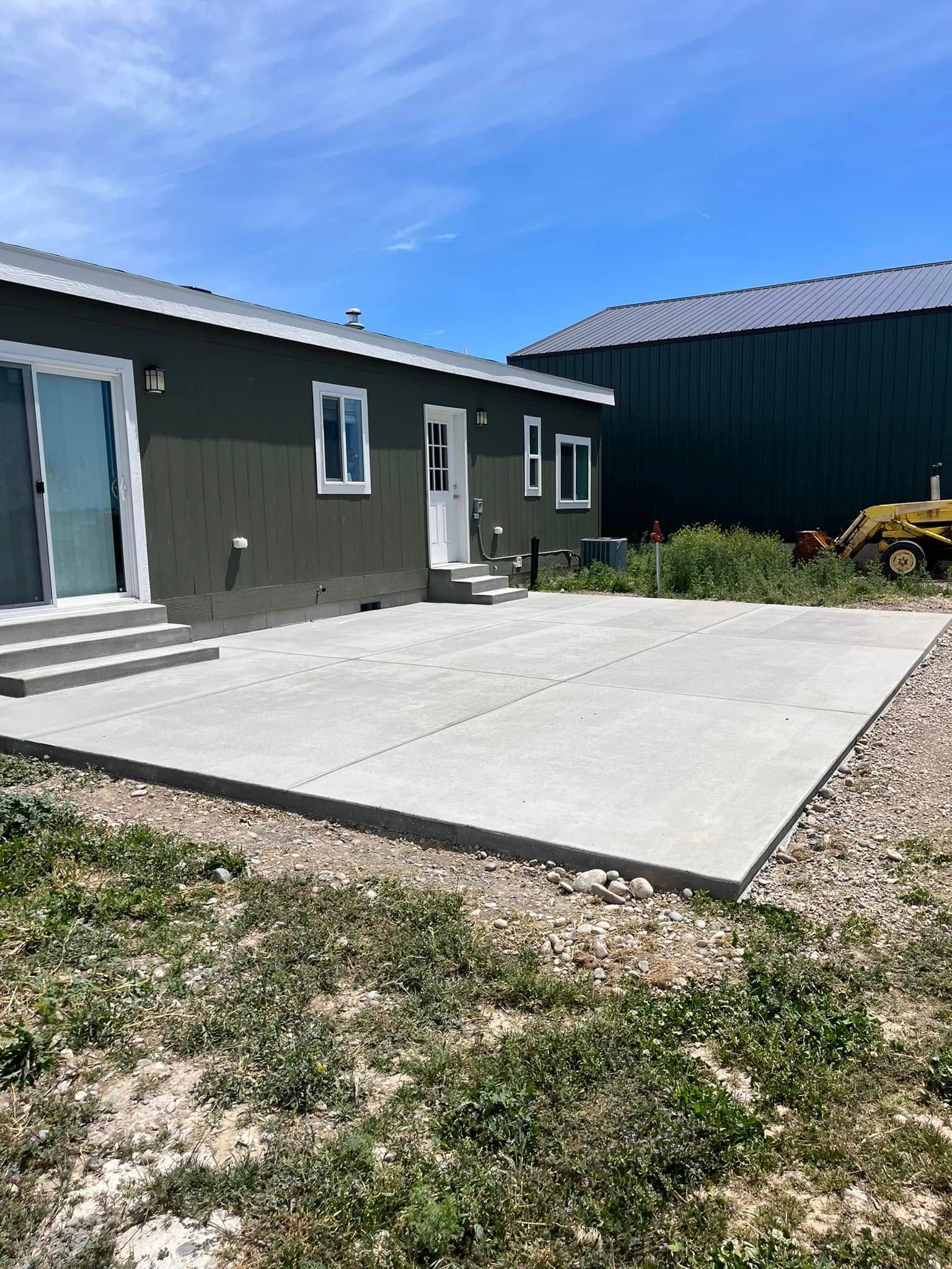 A dark-sided house with a sliding door, a white door, and a new concrete patio slab set against a bright blue sky.