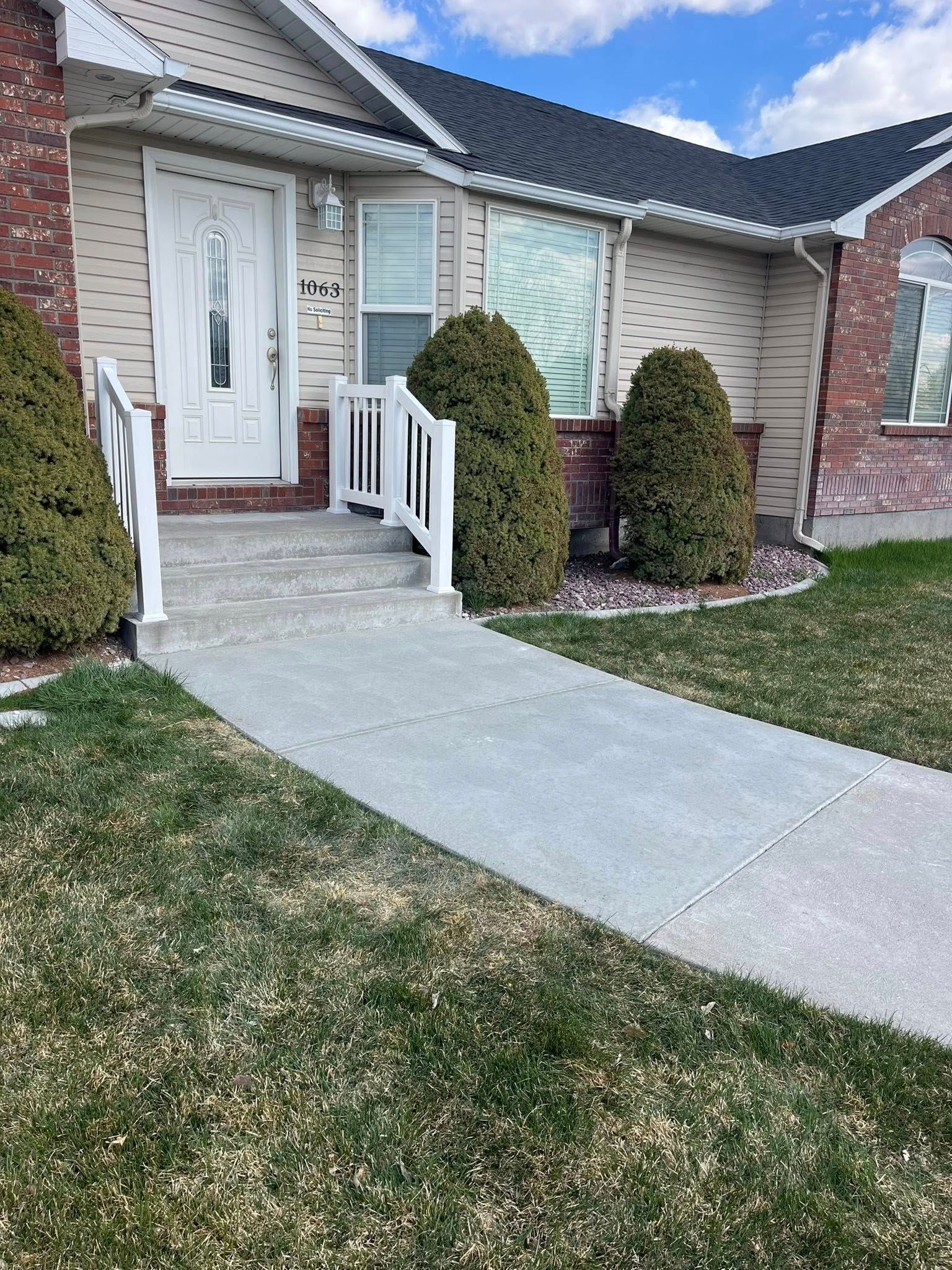 A paved walkway leads to a front door with a white railing and two large evergreen bushes on either side of the entrance.