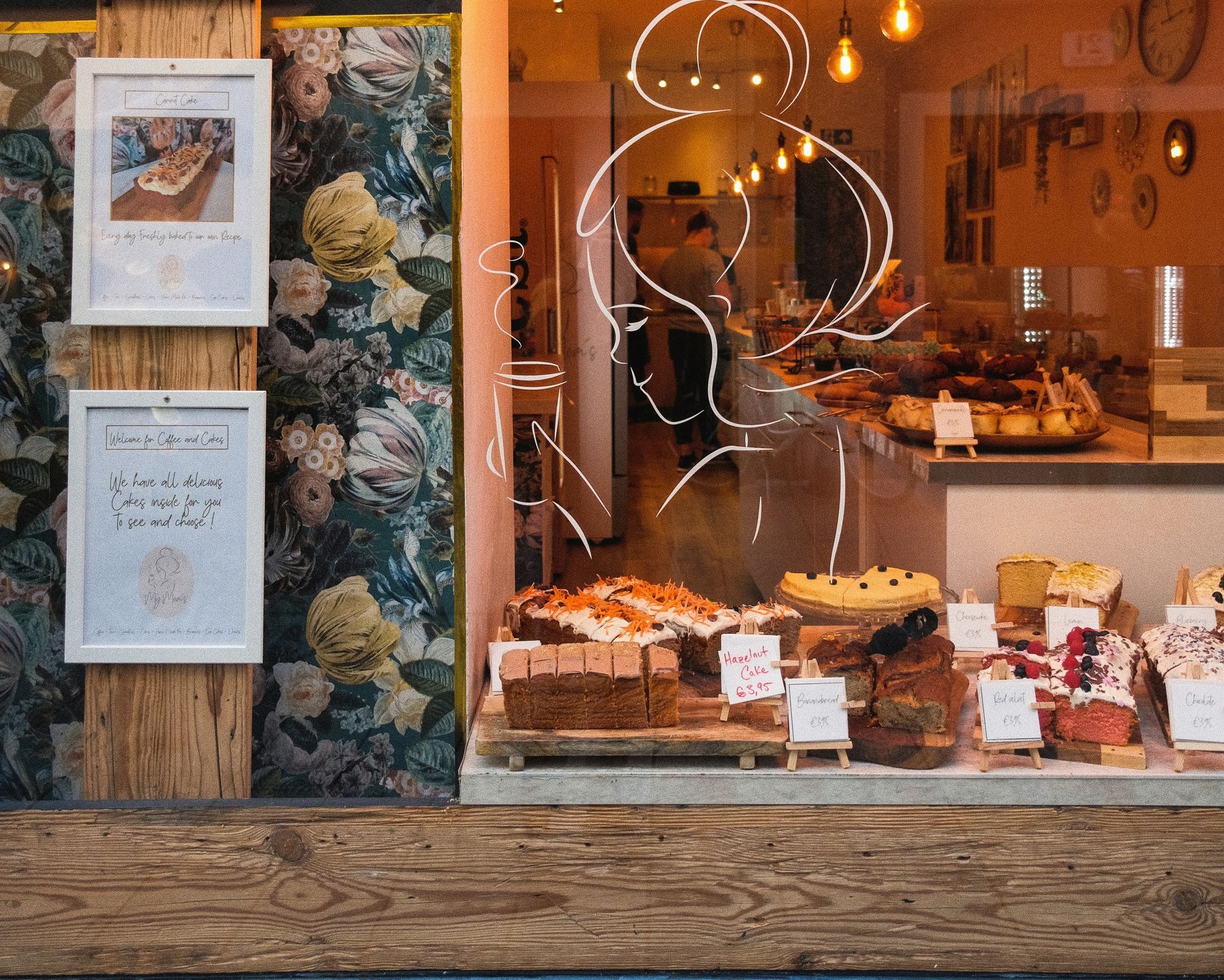 Bakery storefront with cakes, floral wallpaper, and a drawing on the glass.
