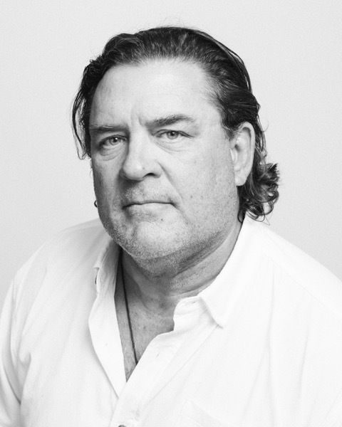A black-and-white portrait of a person with shoulder-length hair wearing a white collared shirt and a necklace.