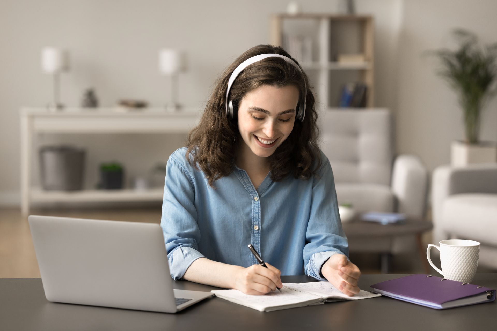 A woman wearing headphones is sitting at a desk with a laptop and writing on a piece of paper.