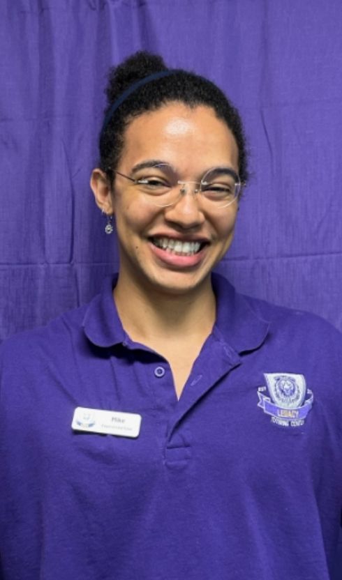 A woman wearing glasses and a purple shirt is smiling in front of a purple curtain.