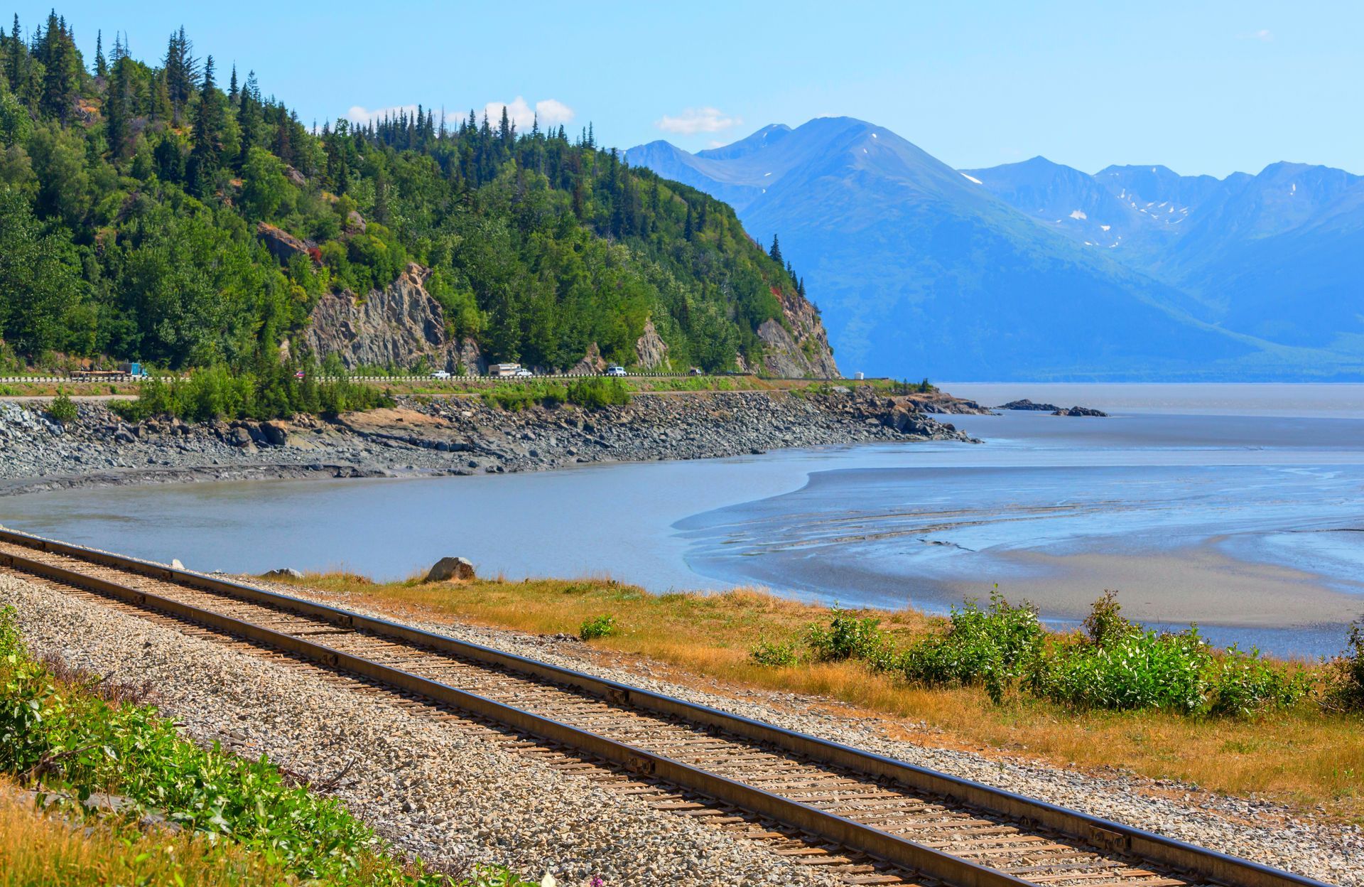 Railroad tracks along a rocky shoreline with forested mountains and calm blue water in the distance