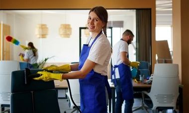 Office cleaners in blue aprons and yellow gloves. Smiling woman cleans, others dust and wipe.