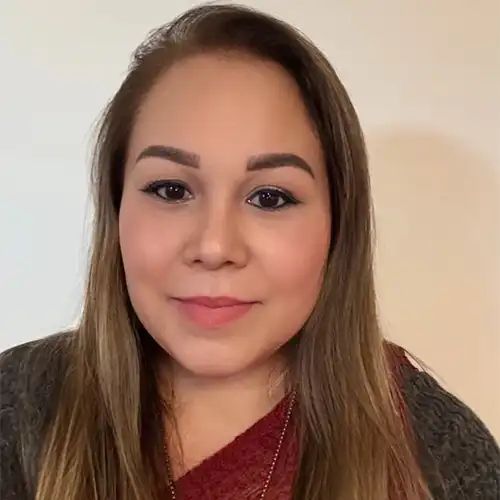 Woman with long brown hair, wearing a gray sweater and red shirt, smiling at the camera.