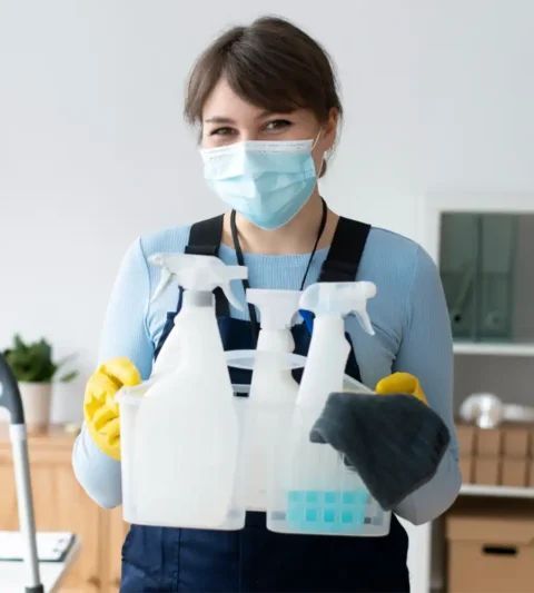 Woman wearing mask and gloves holding cleaning supplies.