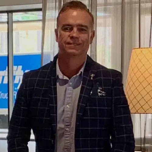 Man in a blue plaid blazer, button-down shirt, smiles at the camera in a building with a window.