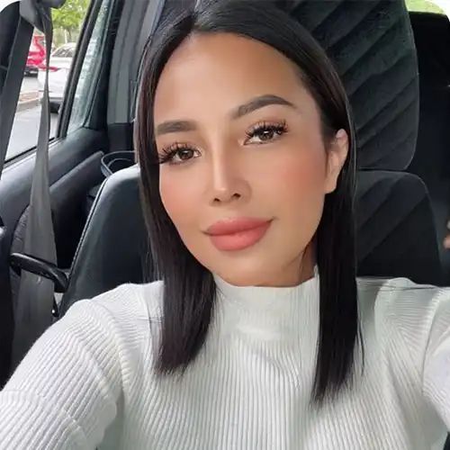 Woman with dark hair in a car, wearing a white sweater, looking at the camera with a neutral expression.