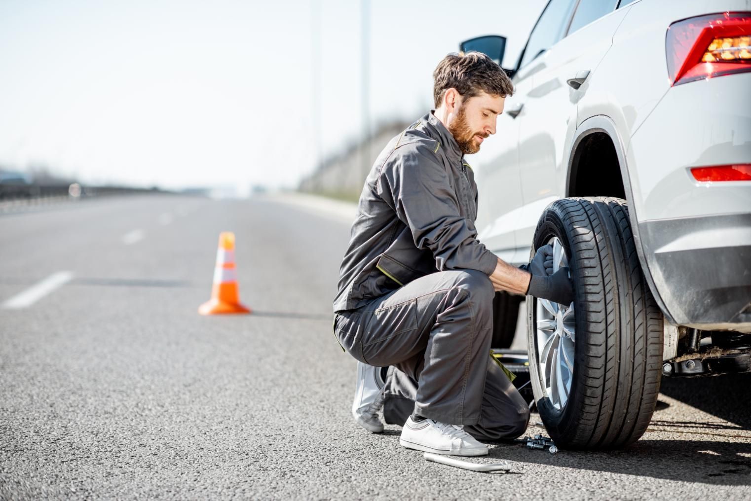 Man changes a car tire on the side of a road, with safety cone visible.