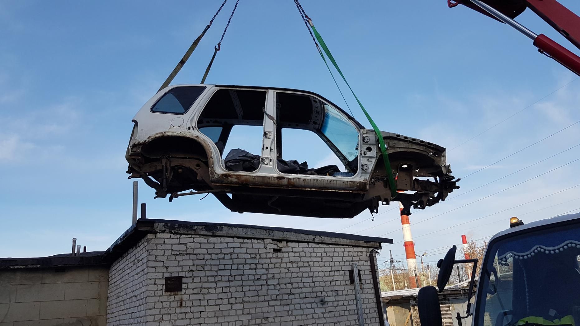 A car frame is hoisted by a crane against a blue sky, near a brick building.