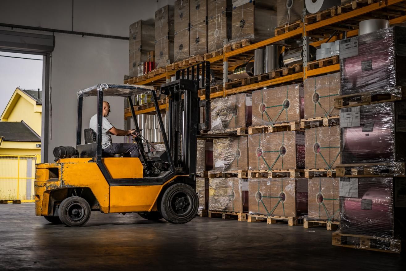 Yellow forklift in a warehouse loading shelves with palletized boxes, driver visible.