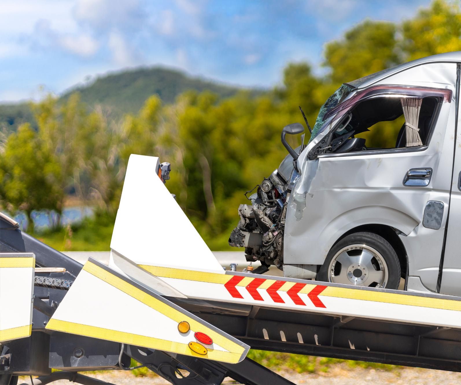 Damaged van on a tow truck, front end crushed. Mountains and trees in the background.