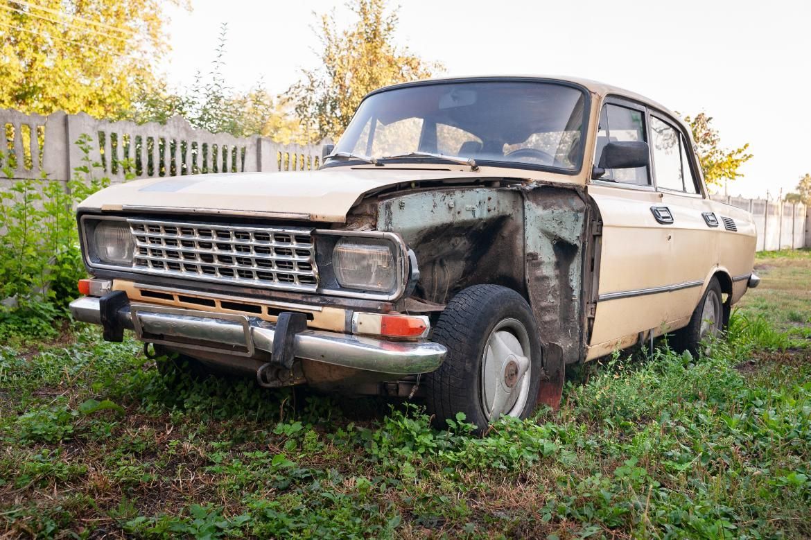 Beige and rust-colored abandoned car missing door and parts, parked in overgrown grass with a fence in the background.
