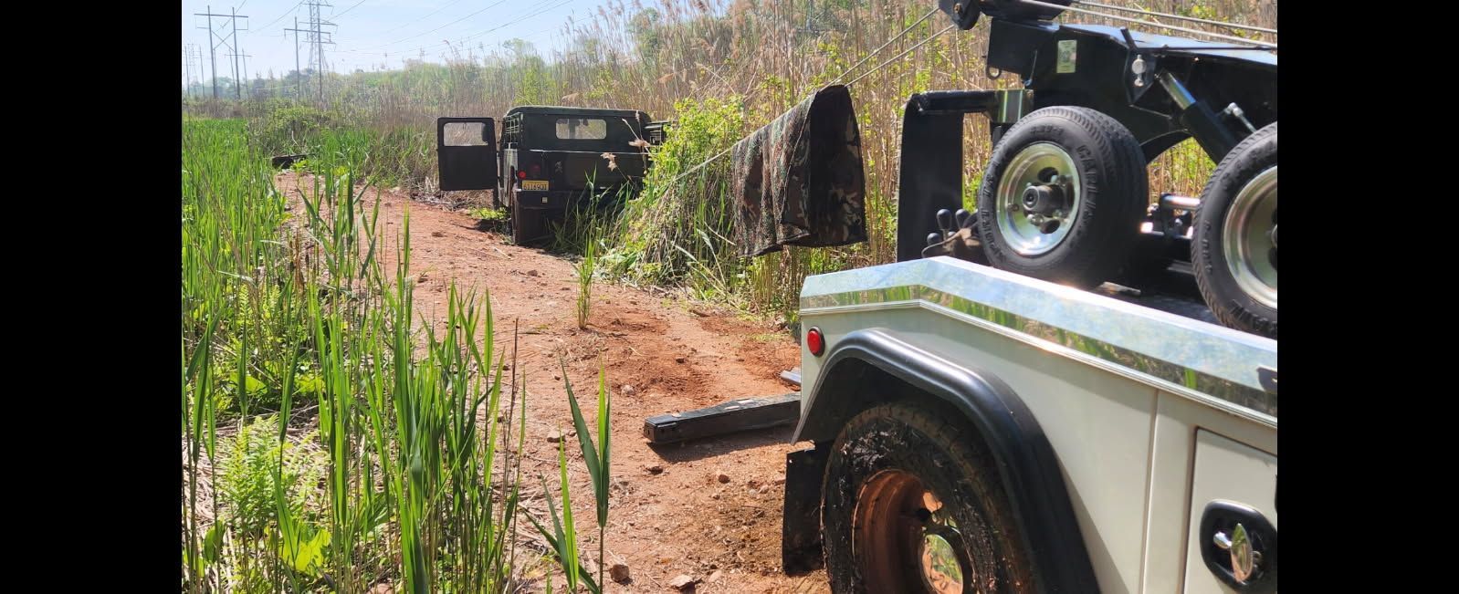 A white tow truck on a dirt path towing a dark vehicle through a grassy area.