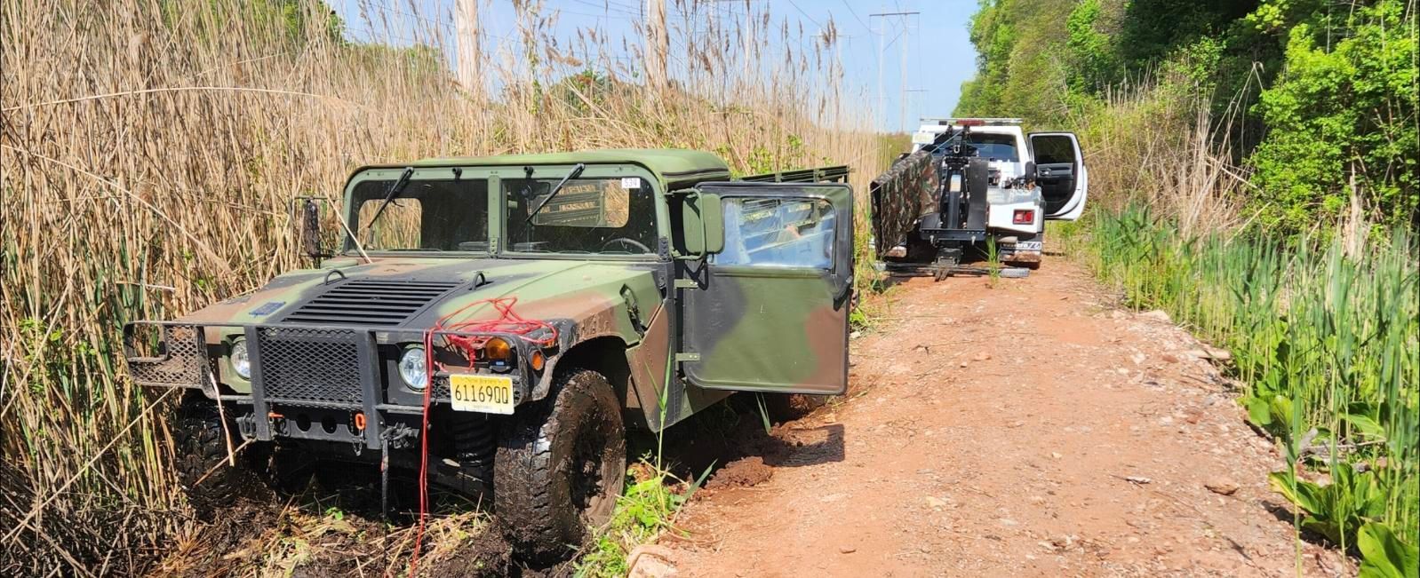 An olive drab Humvee stuck on a muddy road with a white SUV behind it.  Tall grass and foliage on both sides.